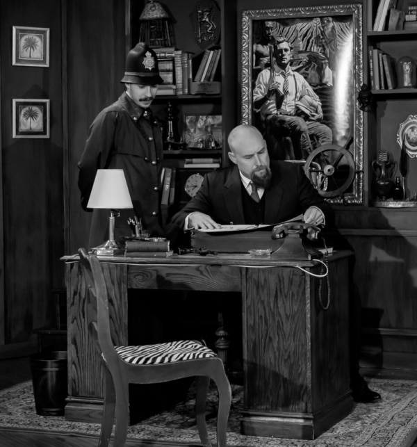 Greyscale photo of Aaron as the Welsh Inspector sitting at a desk looking at a document with another character standing over him